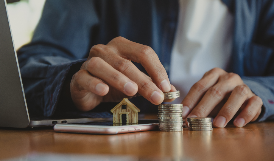 Uma pessoa empilha moedas em uma escrivaninha ao lado de uma pequena casa de madeira, um smartphone e um laptop, simbolizando a poupança ou o investimento em imóveis e considerando o que são passivos em finanças pessoais. O foco está nas mãos que organizam as moedas. - Efacont