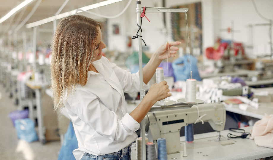 Uma mulher com cabelos loiros na altura dos ombros e camisa branca ajusta um carretel de linha em uma máquina de costura industrial em uma fábrica têxtil de indústria e retalho espaçosa e bem iluminada, repleta de estações de costura e tecidos coloridos ao fundo. - Efacont
