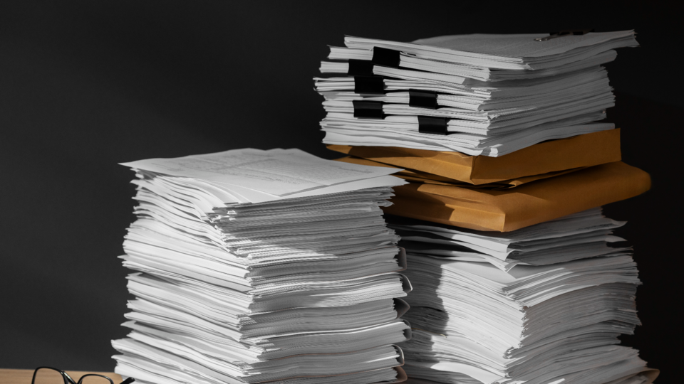 Duas grandes pilhas de documentos em papel repousam sobre uma mesa, alguns cuidadosamente recortados ou grampeados, com dois envelopes pardos no meio de uma pilha — talvez aguardando revisão para medidas de simplificação legislativa por empresas. Um fundo escuro e óculos completam a cena. - Efacont
