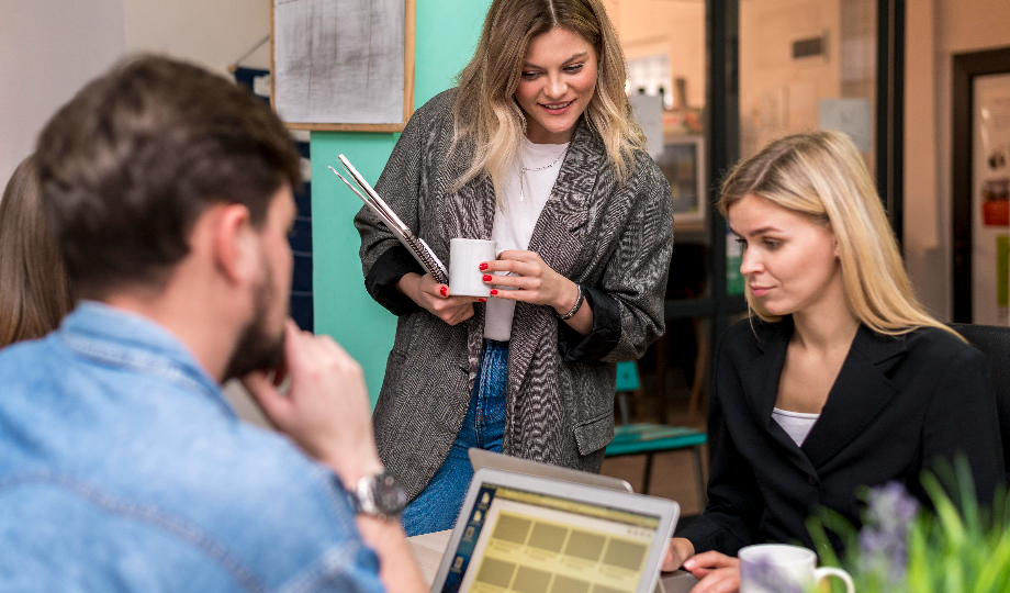 Três pessoas colaboram em um escritório, parecendo envolvidas em uma discussão sobre temas como "o que são as pme". Uma mulher de blazer senta-se em uma mesa com seu laptop, enquanto seus colegas seguram uma caneca e um tablet, sorrindo e olhando para seus computadores. - Efacont