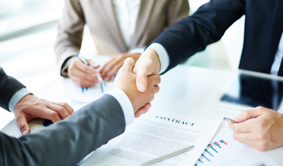 Duas pessoas em ternos de negócios apertam as mãos sobre um contrato e gráficos financeiros em uma mesa, com uma terceira pessoa fazendo anotações. A cena sugere um acordo comercial ou uma negociação bem-sucedida em um ambiente de escritório profissional. - Efacont