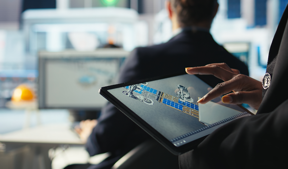 Uma pessoa segura um tablet que exibe um modelo 3D detalhado de um satélite. No fundo desfocado, duas pessoas sentadas em mesas com monitores de computador, refletindo um escritório focado em investimento em automação e inovação. - Efacont