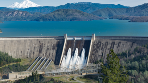 Uma grande represa de concreto libera água por meio de seus vertedouros em um rio abaixo, cercado por colinas e florestas verdejantes - demonstrando resiliência hídrica com um vasto reservatório azul e picos cobertos de neve que se erguem à distância. - Efacont