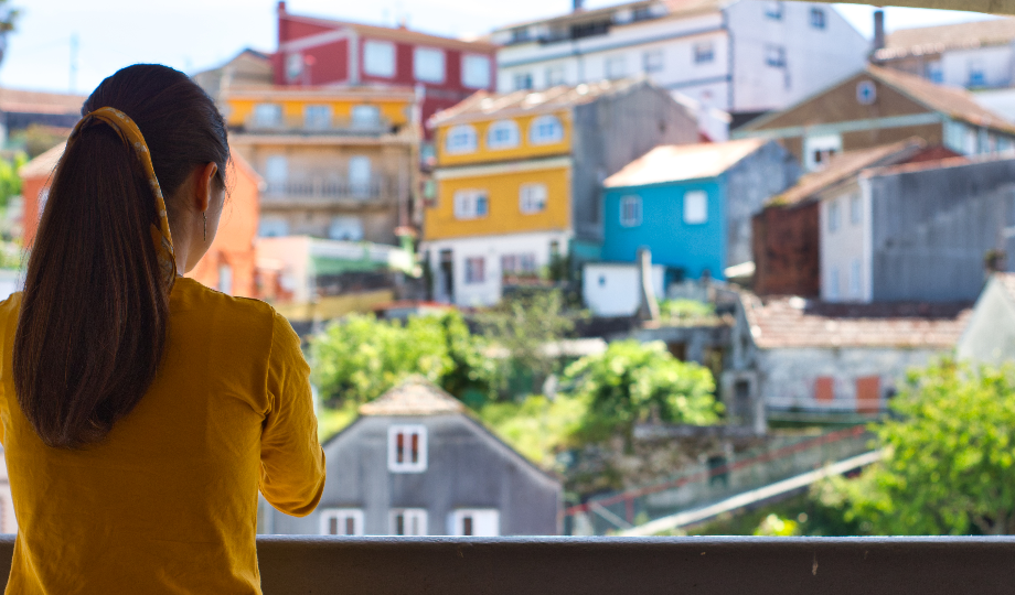 Uma mulher com longos cabelos castanhos em um rabo de cavalo e um top amarelo está em uma varanda, olhando para uma encosta de casas coloridas - uma cena que destaca o charme do turismo em Portugal e sua importância econômica sob o crescimento de 2024. - Efacont