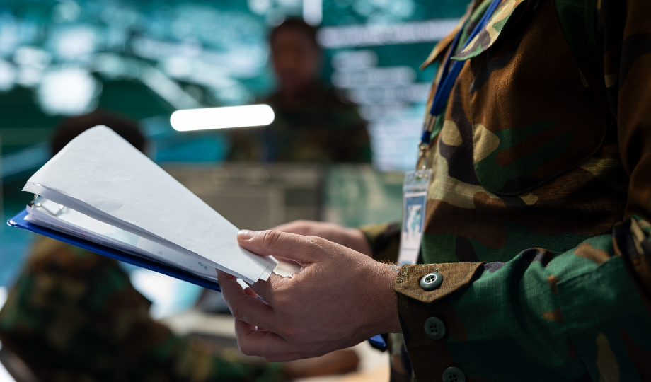 Uma pessoa em uniforme militar camuflado segura uma pasta com papéis, destacando o investimento na defesa nacional. Ao fundo, outros funcionários uniformizados trabalham em computadores em uma sala de controle com telas e informações borradas. - Efacont