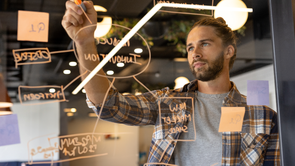 Um homem de barba e cabelos presos escreve em uma parede de vidro cheia de ideias de negócios, diagramas e notas adesivas. Concentrado no planejamento, ele veste uma camisa xadrez sobre uma camiseta cinza em um escritório moderno e bem iluminado com luzes desfocadas. - Efacont