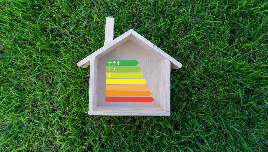 Uma estrutura de madeira em forma de casa fica na grama verde. Em seu interior, um gráfico de classificação de eficiência energética do Programa E-LAR varia de verde (mais eficiente, rotulado como +++) na parte superior a vermelho (menos eficiente, rotulado como -) na parte inferior. - Efacont