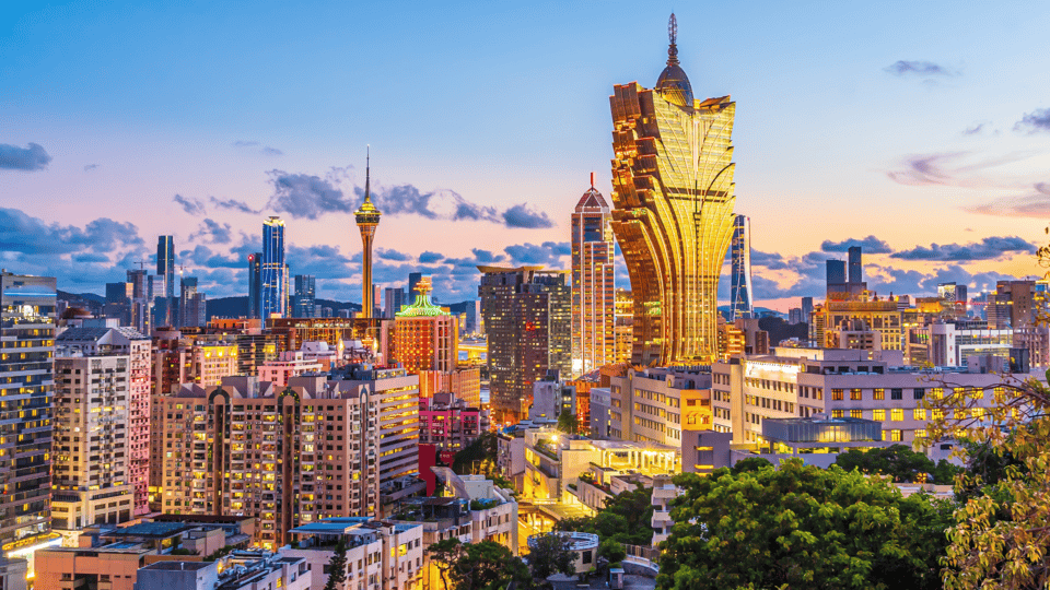 Paisagem urbana de Macau ao anoitecer, com um horizonte de edifícios altos e arranha-céus. O hotel Grand Lisboa se destaca, iluminado acima da cidade que sedia a Feira Internacional de Macau, com céus coloridos à noite e luzes brilhantes da cidade em primeiro plano. - Efacont