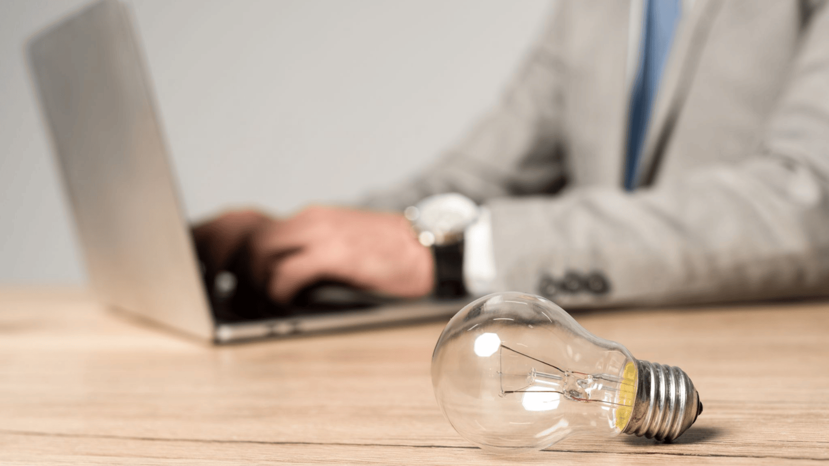 Uma lâmpada clara está sobre uma mesa de madeira em primeiro plano, simbolizando o que é capital intelectual. No plano de fundo desfocado, uma pessoa de terno cinza e gravata azul digita em um laptop, sugerindo inovação em um ambiente de escritório. - Efacont