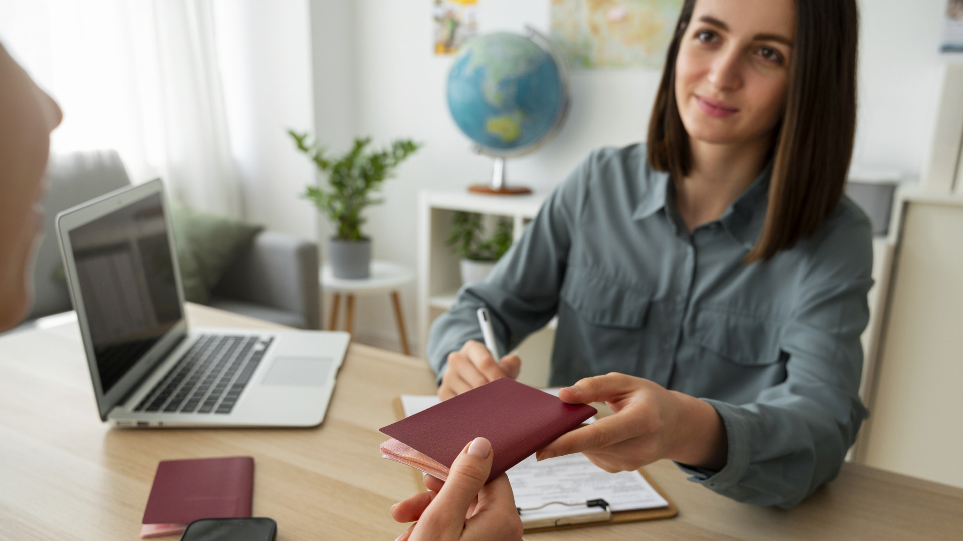 Uma mulher está sentada em uma mesa, entregando um passaporte a outra pessoa enquanto sorri e segura uma caneta. Em sua mesa, ao lado de um laptop e de uma prancheta, há materiais para a Medida de Apoio ao Regresso de Emigrantes a Portugal, além de uma decoração de viagem ao fundo. - Efacont