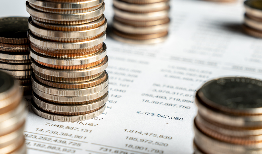 Pilhas de moedas variadas são colocadas em um documento financeiro com números impressos, simbolizando dinheiro e finanças. O fundo desfocado sugere um planejamento financeiro mais amplo, vinculando a cena às discussões sobre o Orçamento do Estado 2026. - Efacont