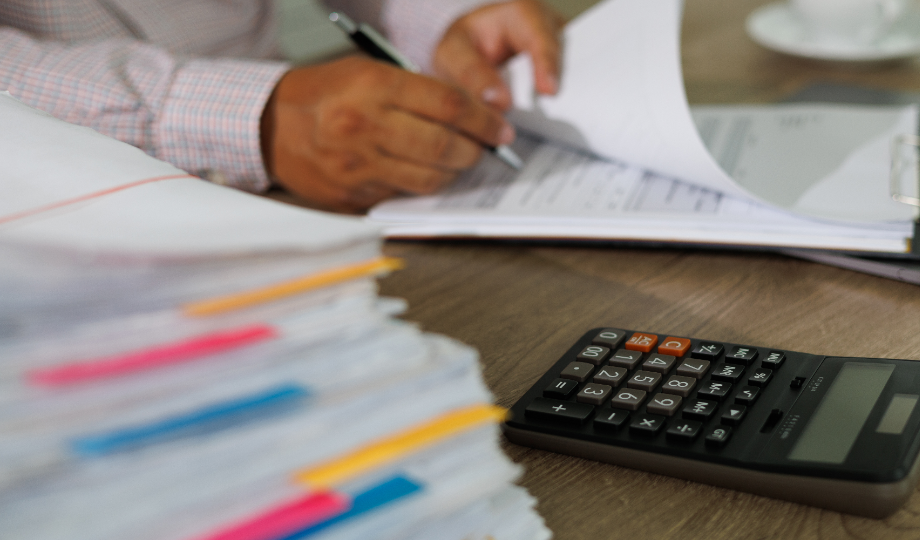 Uma pessoa de camisa listrada revisa e escreve em papéis em uma mesa, possivelmente analisando o Orçamento do Estado 2026. Uma grande pilha de documentos com abas coloridas está em primeiro plano e uma calculadora está próxima, com foco apenas nas mãos e nos documentos. - Efacont