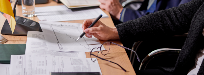 Uma pessoa em trajes de negócios está sentada em uma mesa, segurando uma caneta sobre um gráfico de linhas impresso sobre a constituição de empresas em Portugal. Próximo a ela há óculos, documentos e uma pequena bandeira alemã. Outra pessoa com documentos está parcialmente visível ao fundo. - Efacont