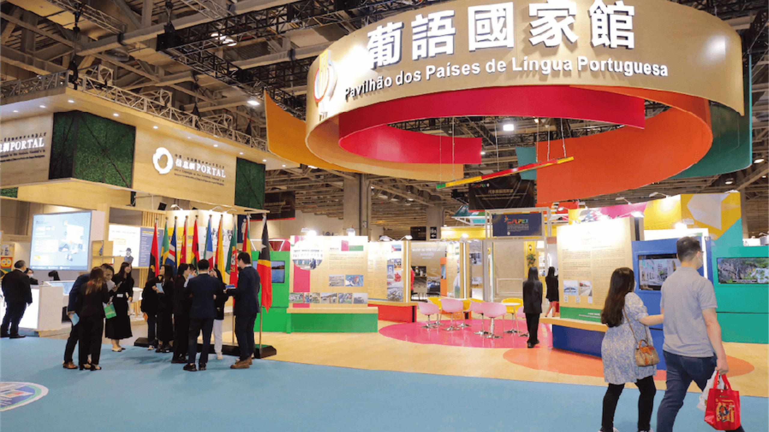 Um colorido salão de exposições na 31.ª Feira Internacional de Macau apresenta estandes sob uma grande placa circular onde se lê "Pavilhão dos Países de Língua Portuguesa". Bandeiras nacionais alinham-se em um estande enquanto os visitantes caminham, conversam e exploram as exposições sob luzes brilhantes. - Efacont