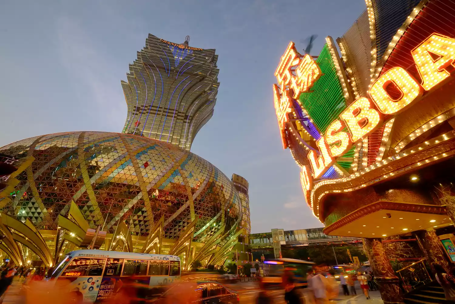 Uma paisagem urbana vibrante ao anoitecer apresenta o Grand Lisboa Hotel and Casino em Macau, com seu design exclusivo e iluminado em forma de lótus. Sinais de neon brilhantes, incluindo um que diz "LISBOA", iluminam a rua movimentada cheia de pessoas e veículos. - Efacont