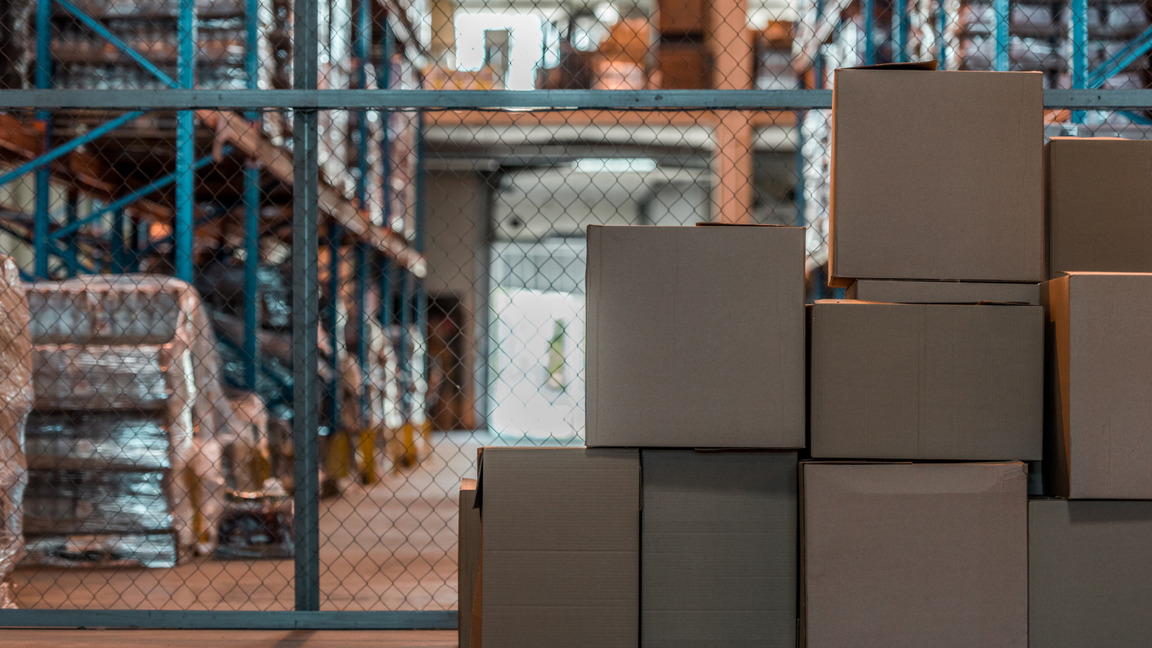 Caixas de papelão empilhadas estão no primeiro plano de um armazém, refletindo a eficiência da Transporte e Logística Moçambique. Estantes de metal repletas de mercadorias embaladas se alinham ao fundo, e uma cerca de arame separa as caixas do restante da área de armazenamento. O espaço parece organizado e industrial. - Efacont