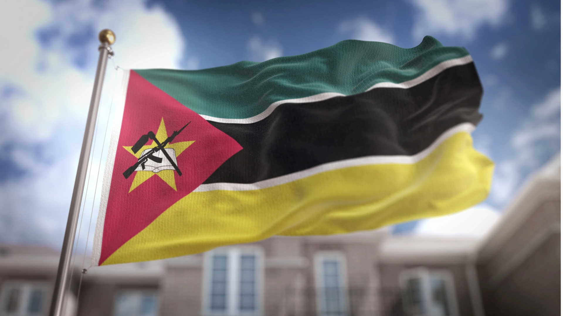 A bandeira de Moçambique está hasteada na Feira Internacional de Maputo. Ela apresenta listras verdes, pretas e amarelas, um triângulo vermelho e uma estrela amarela com um livro, uma enxada e um AK-47. Um céu azul nublado e um prédio em foco difuso aparecem ao fundo. - Efacont