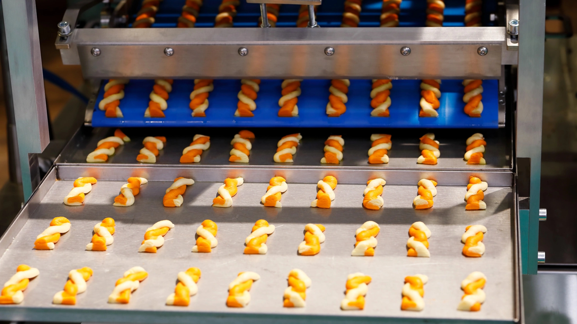 Lanches torcidos, com listras amarelas e brancas, saem de uma máquina e são transportados por uma esteira rolante, dispostos em fileiras organizadas, prontos para o processamento posterior ou para a embalagem em uma moderna unidade de produção alimentar, refletindo o crescimento industrial observado em Moçambique. - Efacont