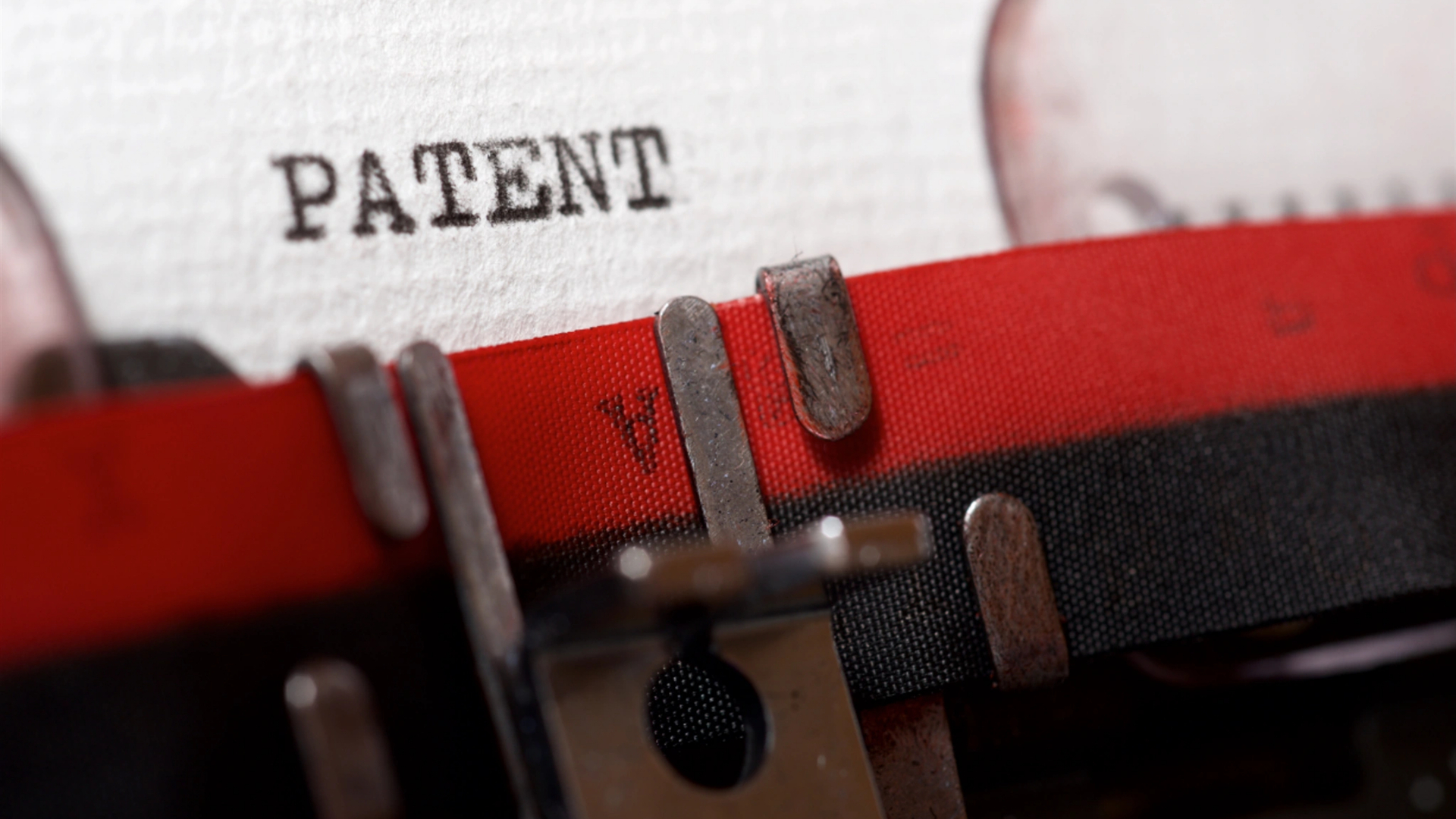 Close-up de uma máquina de escrever vintage digitando a palavra “PATENT” em letras pretas em negrito sobre papel branco texturizado, evocando o processo clássico de registro de patentes. A fita de tinta vermelha e preta e as barras de caracteres metálicas aparecem em primeiro plano. - Efacont